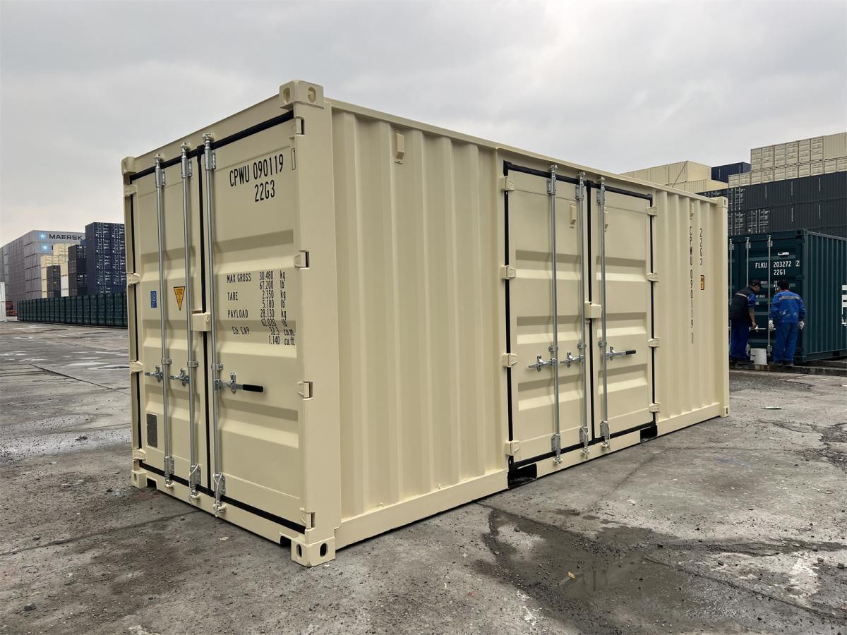 A three-quarter perspective view of a closed ivory shipping container on a concrete lot. This angle shows both the standard rear cargo doors and a second set of double doors integrated into the long side panel. Other stacked shipping containers are visible in the background under an overcast sky.