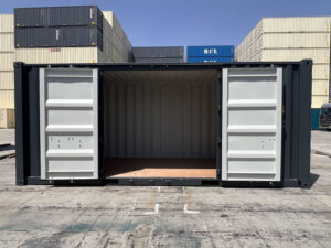 A wide side view of the dark gray shipping container with its side double doors propped completely open. The interior features a clean plywood floor and light gray internal walls, with various cream and blue shipping containers stacked in the background under a clear blue sky.