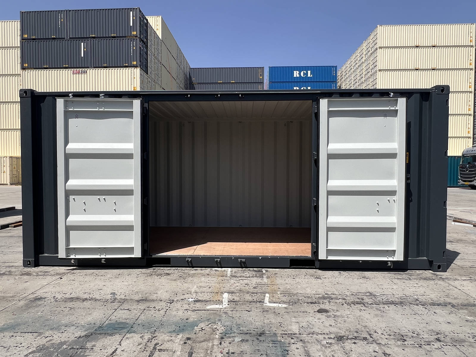A wide side view of the dark gray shipping container with its side double doors propped completely open. The interior features a clean plywood floor and light gray internal walls, with various cream and blue shipping containers stacked in the background under a clear blue sky.