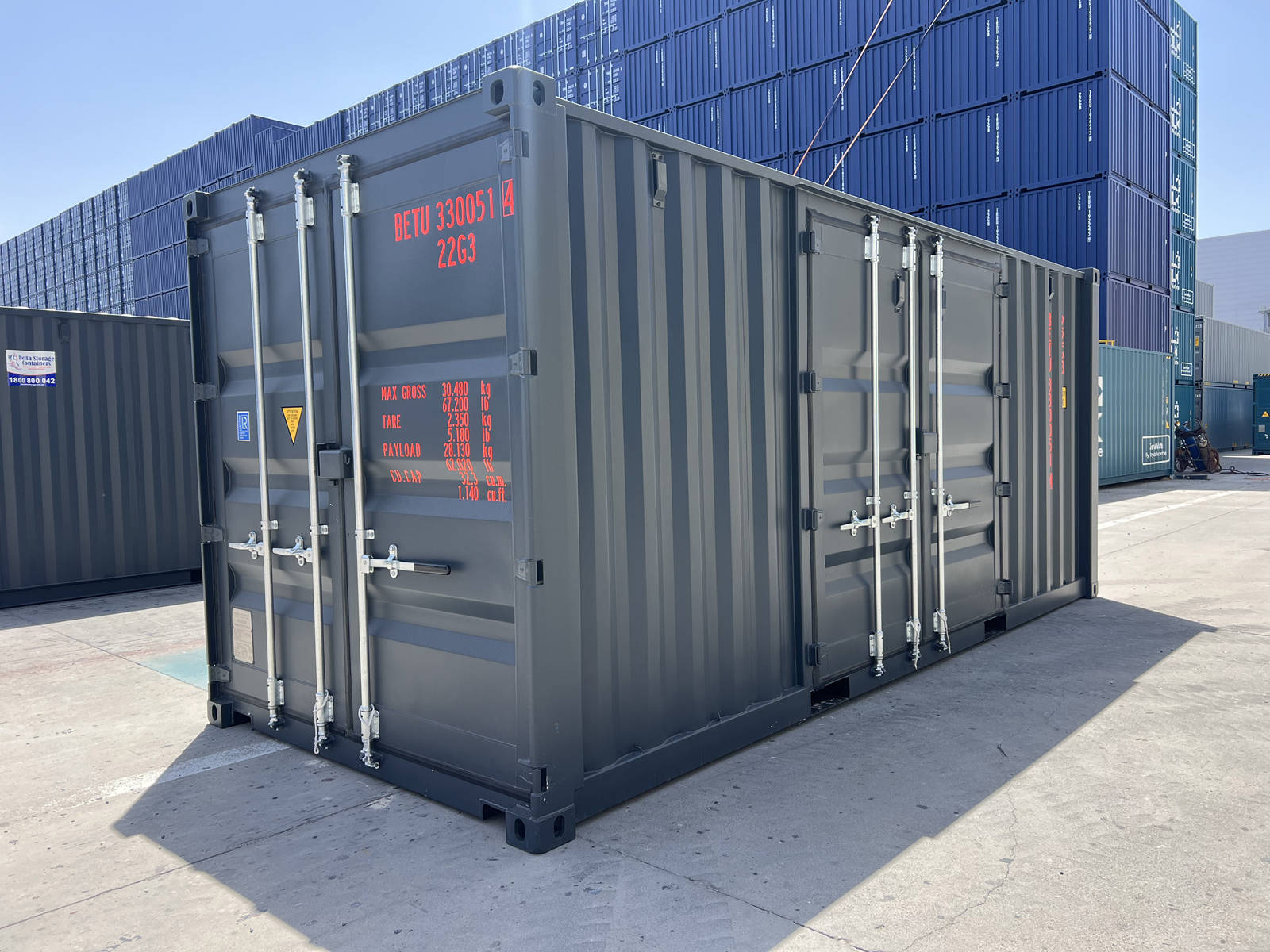 A three-quarter perspective view of the dark gray shipping container on a bright, concrete terminal. The shot displays the standard end doors with orange lettering alongside an additional set of double doors built into the corrugated side panel. Stacks of blue containers rise in the background.